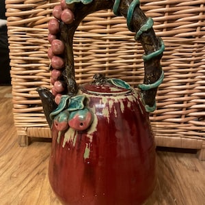 Könnte beinhalten: Eine Keramik-Teekanne mit tiefrotem Korpus und gebogenem Henkel. Der Henkel ist braun mit grünen, rankenartigen Akzenten. Die Teekanne ist mit rosa Früchten und grünen Blättern verziert. Ausguss und Deckel sind dunkelbraun.