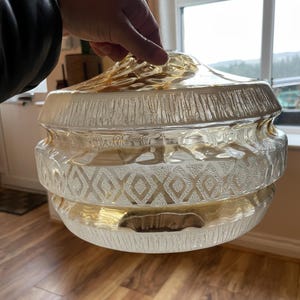 Puede incluir: Una lámpara colgante vintage de cristal transparente con la parte superior e inferior doradas. El cristal tiene un diseño texturizado y acanalado en la parte superior e inferior, con un patrón de diamantes en el medio. La lámpara está sujeta por una mano.