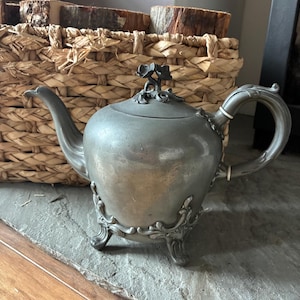 May include: A metallic grey teapot featuring a curved spout, a handle with two white rings, and three ornate feet. The lid is topped with a sculpted floral finial. The body of the teapot has a smooth, matte finish with decorative scrollwork at the base.