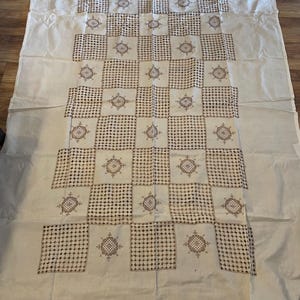 Pode incluir: Toalha de mesa creme com um padrão geométrico. O design apresenta quadrados alternados de bordado pontilhado e floral em um tom marrom. A toalha de mesa é retangular e parece ser feita de um tecido macio.