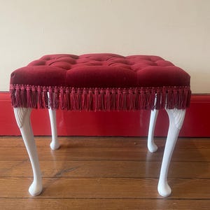 May include: A small, tufted ottoman with a deep red velvet cushion and white, curved legs. The cushion is trimmed with matching red fringe. The legs are painted white, creating a contrast with the rich red of the cushion.