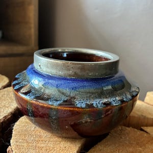 Op de afbeelding: Een kleine, ronde keramische kom met een metallic zilveren rand. Het lichaam van de kom heeft een glanzend glazuur met een diepbruine basiskleur, die overgaat in een levendige blauwe band bij de bovenkant, geaccentueerd door textuurrijke, witte druipsporen.