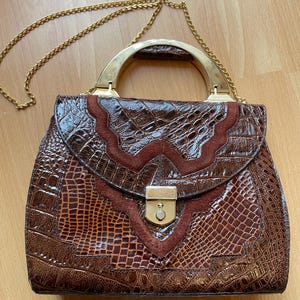 May include: A vintage brown handbag with a gold-tone handle and chain strap. The bag features a crocodile-textured pattern with a suede accent on the flap. The clasp is gold-toned.