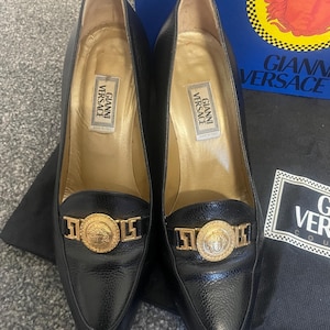 Może przedstawiać: Para czarnych sk&oacute;rzanych szpilek ze szpiczastymi noskami. Każdy but posiada złoty medalion z greckim wzorem głowy i nazwą marki "Gianni Versace". Obcasy mają wysokość około 7,6 cm.