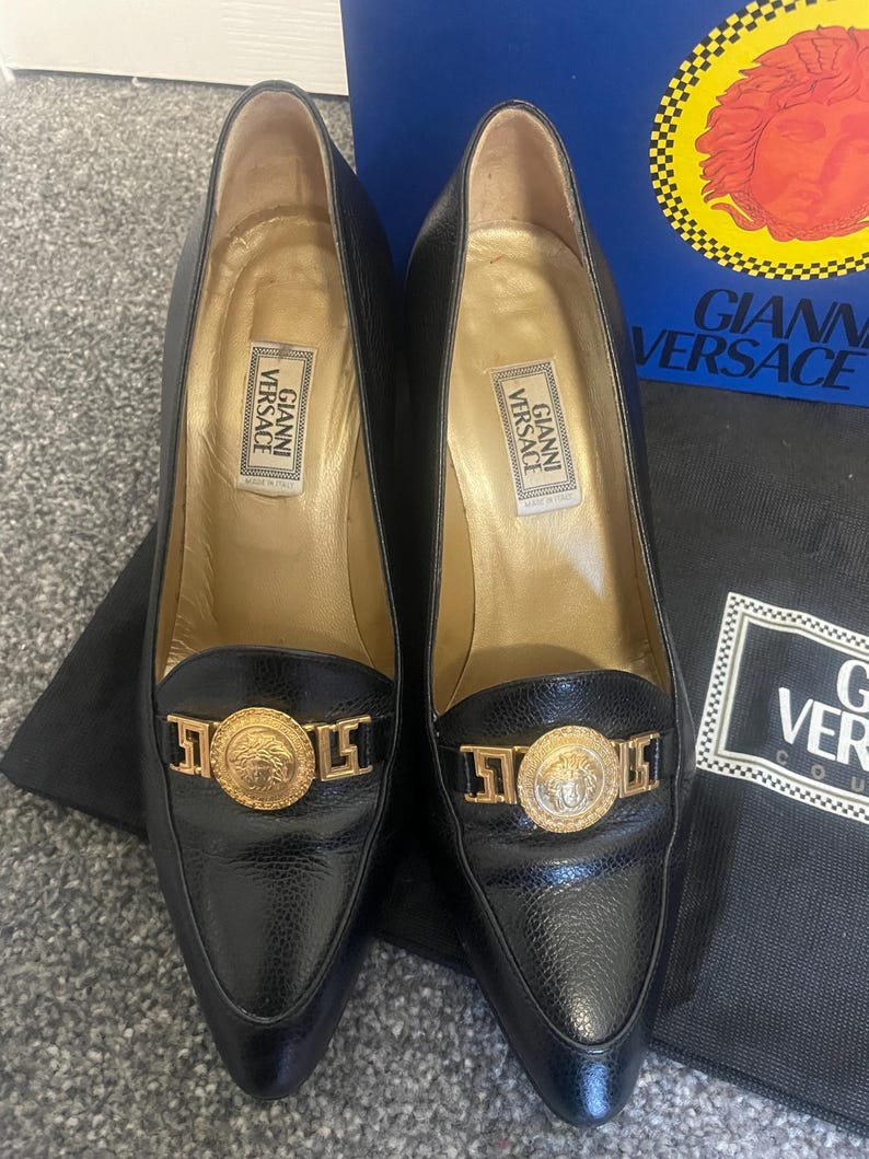 Może przedstawiać: Para czarnych sk&oacute;rzanych szpilek ze szpiczastymi noskami. Każdy but posiada złoty medalion z greckim wzorem głowy i nazwą marki "Gianni Versace". Obcasy mają wysokość około 7,6 cm.