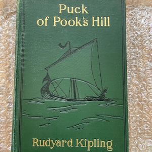 Puede incluir: Un libro de tapa dura verde con letras doradas titulado "Puck of Pook's Hill" de Rudyard Kipling. La portada presenta una ilustración en blanco y negro de un barco navegando en el agua.