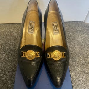Może przedstawiać: Para czarnych sk&oacute;rzanych szpilek Gianni Versace ze złotymi akcentami. Buty mają szpiczaste noski i złoty emblemat na cholewce. Wnętrze jest złote z etykietą Gianni Versace.