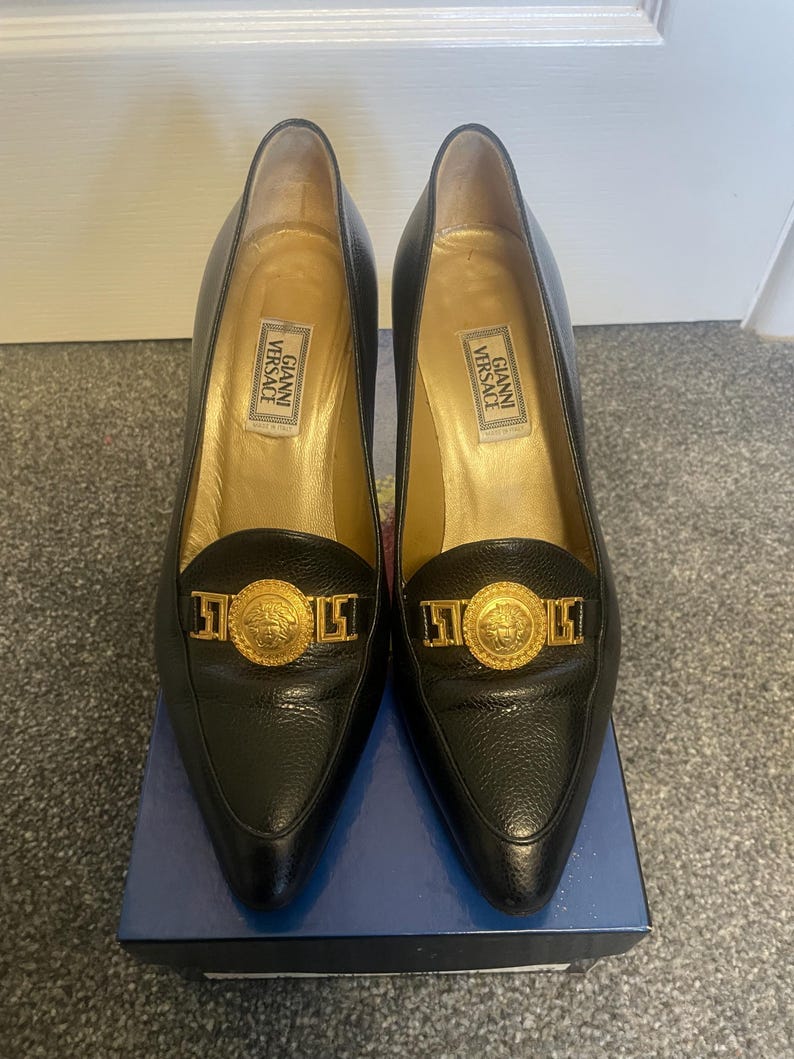Może przedstawiać: Para czarnych sk&oacute;rzanych szpilek Gianni Versace ze złotymi akcentami. Buty mają szpiczaste noski i złoty emblemat na cholewce. Wnętrze jest złote z etykietą Gianni Versace.