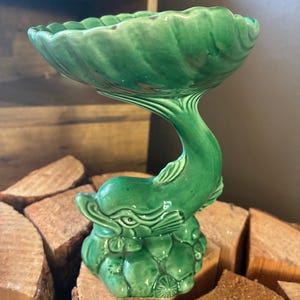 May include: A glossy emerald green ceramic dish featuring a sculpted dolphin base. The dolphin is depicted leaping upward, with its tail supporting a wide, scalloped-edge bowl. The base is textured with detailed coral and sea life elements.