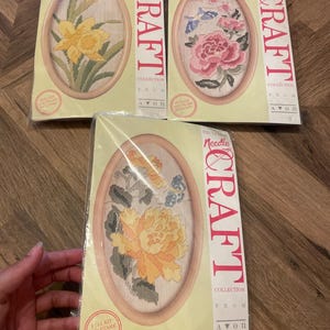 May include: Three sealed needlepoint craft kits from the Avon Exclusive Needlecraft Collection. Each kit features an oval frame and a floral design, including yellow daffodils, pink peonies with a blue butterfly, and orange flowers with green leaves.
