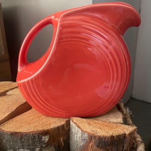 May include: A bright coral-colored ceramic pitcher featuring a rounded body with concentric circular ridges, a curved handle, and a wide pouring spout. The pitcher has a smooth, glossy finish and a distinct, symmetrical design.