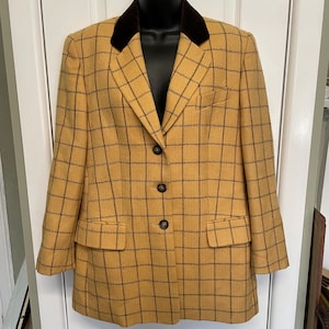 May include: A mustard yellow blazer featuring a dark brown windowpane check pattern. The jacket includes a dark brown velvet collar, three front buttons, two flap pockets at the waist, and one chest pocket on the left side, displayed on a hanger.