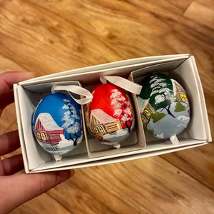 May include: A set of three decorative egg ornaments in a white cardboard box. Each egg features a hand-painted winter landscape with a small house, snow-covered trees, and white glitter accents. Each egg has a white ribbon loop for hanging.