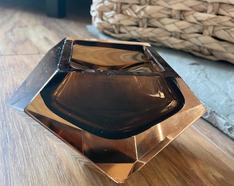 May include: A geometric, brown glass ashtray with a copper-colored rim. The ashtray has a diamond shape with a square opening. The glass is transparent, allowing light to pass through. The ashtray is on a wooden surface.