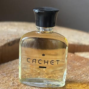 May include: A small, clear glass perfume bottle with a black cap. The word "CACHET" is printed in black on the front, along with a small crown symbol. The bottle is filled with a light-coloured liquid.