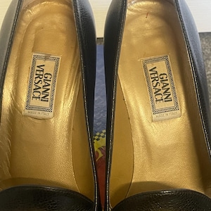 Może przedstawiać: Para czarnych sk&oacute;rzanych but&oacute;w na wysokim obcasie ze złotymi detalami. Buty posiadają złoty emblemat z głową Meduzy i etykietę "GIANNI VERSACE MADE IN ITALY" wewnątrz. Obcasy mają około 7.6 cm wysokości.