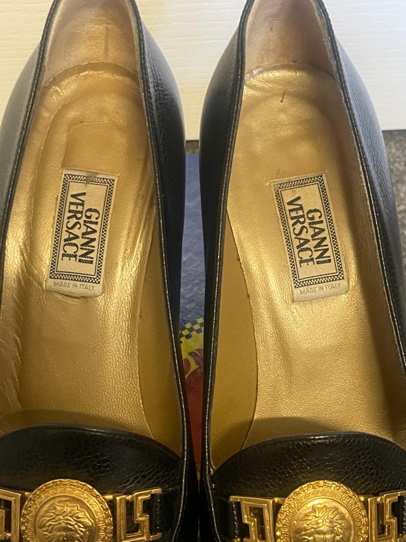 Może przedstawiać: Para czarnych sk&oacute;rzanych but&oacute;w na wysokim obcasie ze złotymi detalami. Buty posiadają złoty emblemat z głową Meduzy i etykietę "GIANNI VERSACE MADE IN ITALY" wewnątrz. Obcasy mają około 7.6 cm wysokości.