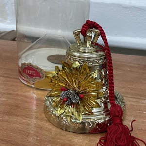 May include: A silver-colored bell with a gold floral accent and a red tassel. The bell has a decorative design and a crown-like top. A clear plastic container is in the background.