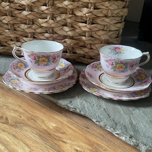 Può includere: Due tazze e piattini abbinati con un motivo floreale rosa. Le tazze hanno un interno bianco e finiture dorate. I piattini sono rosa con bordo dorato e motivo floreale. Il set è posto su una superficie di legno.