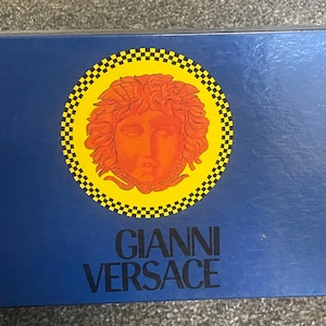 Może przedstawiać: Granatowe pudełko z logo Gianni Versace. Logo zawiera okrągły wz&oacute;r z ż&oacute;łtą obw&oacute;dką i czarno-białym wzorem w szachownicę. Wewnątrz okręgu znajduje się szczeg&oacute;łowa pomarańczowa twarz.