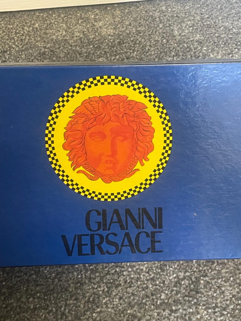Może przedstawiać: Granatowe pudełko z logo Gianni Versace. Logo zawiera okrągły wz&oacute;r z ż&oacute;łtą obw&oacute;dką i czarno-białym wzorem w szachownicę. Wewnątrz okręgu znajduje się szczeg&oacute;łowa pomarańczowa twarz.