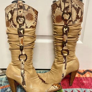 Könnte beinhalten: Ein Paar kniehohe Stiefel mit einem beigefarbenen Lederschaft im gerafften Design, einer spitzen Zehenpartie und einem hohen Holzabsatz. Die Stiefel sind mit braun-cremefarbenen Schlangenmuster-Einsätzen, Metallringen und Zierschnallen versehen.