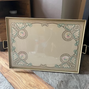 Op de afbeelding: Rechthoekig dienblad met een goudkleurig frame en handgrepen. De lade heeft een crèmekleurig midden met geborduurde bloemen- en bladerontwerpen in roze, groen en grijs. Waarschijnlijk een vintage of antiek item.