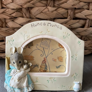 Pode incluir: Uma moldura de foto arqueada na cor creme com o texto Hunca Munca no topo. Uma figura de rato esculpida com um vestido azul segura um rato menor no lado esquerdo. A moldura inclui um inserto amarelo com gráficos de The World of Beatrix Potter.