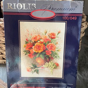 May include: A cross-stitch kit with a floral design of orange and red roses in a vase, and green grapes. The box is dark blue with the text "RIOLIS Premium" and "Tea Roses".