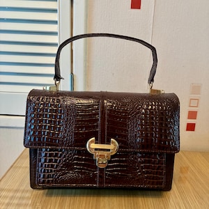 Puede incluir: Un bolso de mano marrón oscuro con una textura brillante de relieve de cocodrilo. El bolso cuenta con un asa superior, una forma rectangular estructurada y un cierre metálico de tono dorado centrado en la solapa frontal.