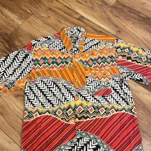 Puede incluir: Una camisa de manga corta con un vibrante estampado geométrico. La camisa presenta una mezcla de colores que incluyen naranja, rojo, negro, blanco y verde. El diseño incluye zigzags, rayas y formas de diamante.