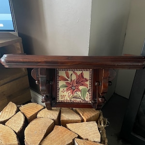 May include: A dark brown wooden wall shelf featuring a central square ceramic tile decorated with a floral illustration of a red lily. The shelf has a horizontal top ledge and decorative carved side supports with rounded wooden accents.