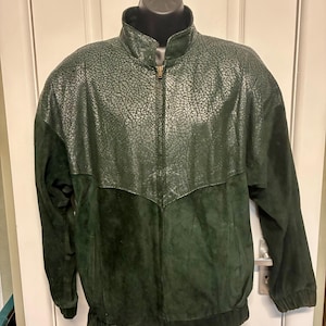 May include: A dark green jacket featuring a textured, cracked-pattern leather upper panel and a smooth suede lower section. The jacket includes a full-length front zip, a stand-up collar, and elasticated cuffs and hem for a structured fit.