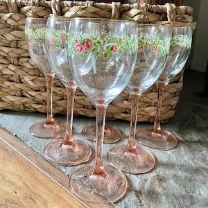 May include: A set of six vintage wine glasses. Each glass has a pink stem and base, with a clear bowl adorned with a floral garland of pink roses and green foliage. The glasses are displayed in front of a wicker basket.