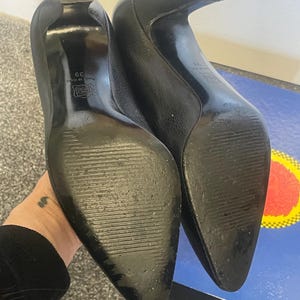 Może przedstawiać: Para czarnych sk&oacute;rzanych szpilek z teksturowanym wykończeniem. Buty mają spiczasty nosek i zakrzywiony obcas. Widoczne są podeszwy, z rozmiarem 39 i wzorem. Obcasy mają około 7.6 cm wysokości.