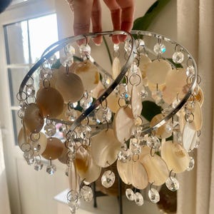 May include: A circular chrome light fixture frame adorned with cascading strands of iridescent, round, flat shell discs and clear, faceted glass beads. The structure features two intersecting metal rings that support the hanging decorative elements.
