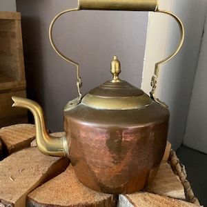 May include: A copper tea kettle featuring a hammered texture on the body, a brass spout, a brass lid with a central finial, and a large arched brass handle. The kettle has a rounded base and a polished finish that reflects light across its surface.