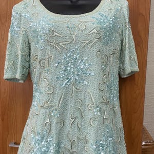 May include: A pale green, short-sleeved top adorned with detailed sequin and bead work. The top has a round neckline and a floral pattern with light blue sequins. The fabric has a slight sheen, enhancing its elegant look. The top is displayed on a black mannequin.