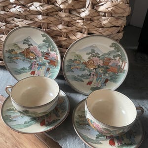 May include: A vintage tea set featuring two teacups, saucers, and plates. The set has a cream-colored base with a landscape design in shades of green, pink, and blue. The plates and saucers have a gold trim. The background includes a woven basket and wood.