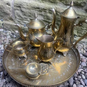 May include: A vintage brass tea set, including a teapot, creamer, sugar bowl, and tea strainer, arranged on a decorative tray. The set has an ornate design and is displayed on a bed of pebbles.
