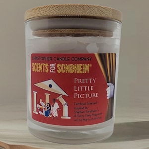 May include: A glass candle jar with a wooden lid, labeled "Christopher Candle Company SCENTS of SONDHEIM" and "PRETTY LITTLE PICTURE." A wooden spoon and a small puddle of liquid are in the foreground. The candle has a red label with a theater-themed design.