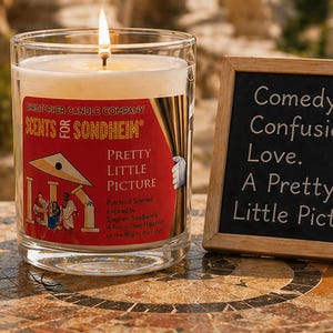 May include: A lit candle in a clear glass jar with a red label that reads "SCENTS of SONDHEIM PRETTY LITTLE PICTURE." Next to it is a small chalkboard with the words "Comedy. Confusion. Love. A Pretty Little Picture."