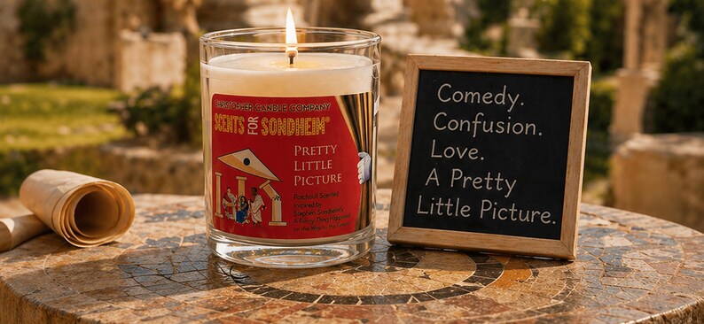 May include: A lit candle in a clear glass jar with a red label that reads "SCENTS of SONDHEIM PRETTY LITTLE PICTURE." Next to it is a small chalkboard with the words "Comedy. Confusion. Love. A Pretty Little Picture."