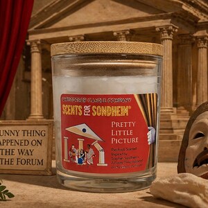 May include: A glass candle jar with a wooden lid, labeled "SCENTS FOR SONDHEIM" and "PRETTY LITTLE PICTURE." The label features a red background with a theater-themed illustration. A small framed sign reads "A FUNNY THING HAPPENED ON THE WAY TO THE FORUM."