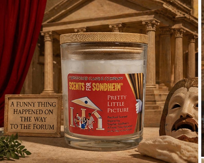 May include: A glass candle jar with a wooden lid, labeled "SCENTS FOR SONDHEIM" and "PRETTY LITTLE PICTURE." The label features a red background with a theater-themed illustration. A small framed sign reads "A FUNNY THING HAPPENED ON THE WAY TO THE FORUM."