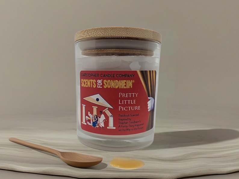 May include: A glass candle jar with a wooden lid, labeled "Christopher Candle Company SCENTS of SONDHEIM" and "PRETTY LITTLE PICTURE." A wooden spoon and a small puddle of liquid are in the foreground. The candle has a red label with a theater-themed design.