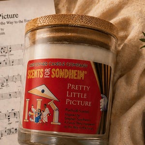 May include: A candle in a glass jar with a bamboo lid, labeled "Scents for Sondheim Pretty Little Picture." The label is red with a theater scene illustration. Sheet music and a golden theater mask are in the background.