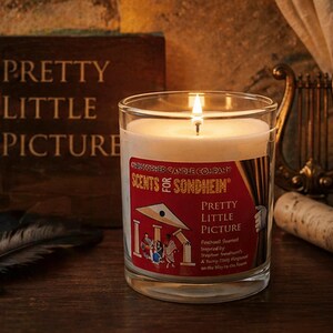 May include: A lit candle in a clear glass jar with a label that reads "Pretty Little Picture." The label also says "Scents for Sondheim." A book with the words "Pretty Little Picture" is in the background.
