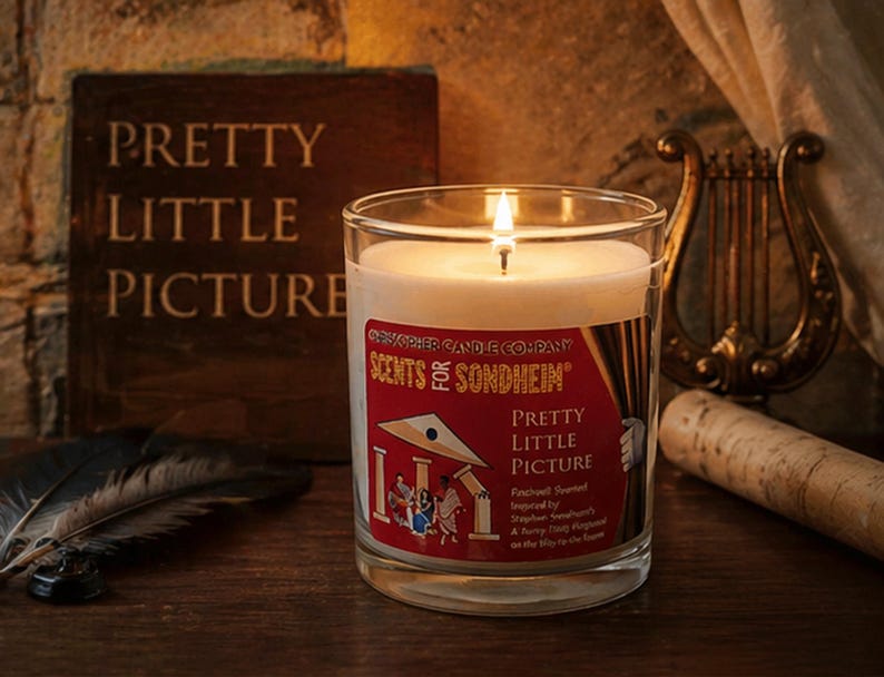 May include: A lit candle in a clear glass jar with a label that reads "Pretty Little Picture." The label also says "Scents for Sondheim." A book with the words "Pretty Little Picture" is in the background.