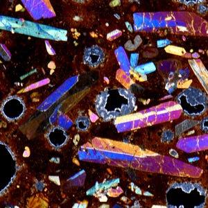 Könnte beinhalten: Ein Dünnschliff von Vulkangestein unter polarisiertem Licht, der eine dunkelbraune Matrix mit irisierenden, vielfarbigen Kristallstrukturen sowie mehreren kreisförmigen, schwarzen, hohlen Blasen in verschiedenen Größen zeigt.